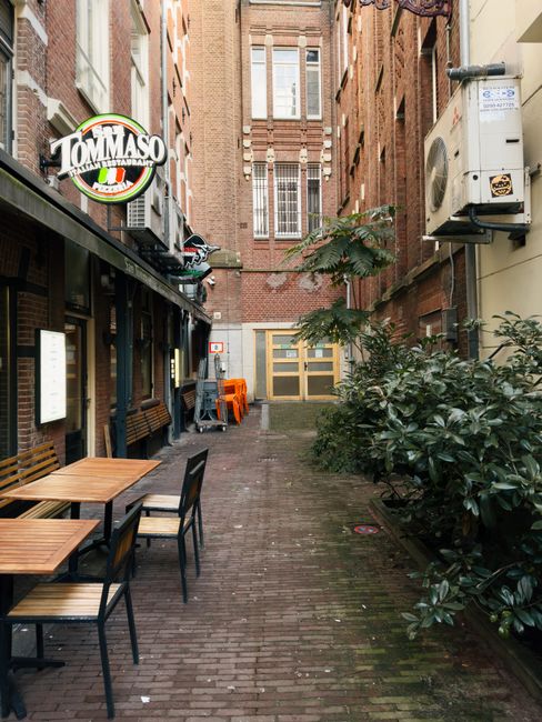 A photograph taken from the ground level shows a narrow street in Amsterdam with tall, closely positioned buildings on either side. The building on the left features a red brick facade with multiple w